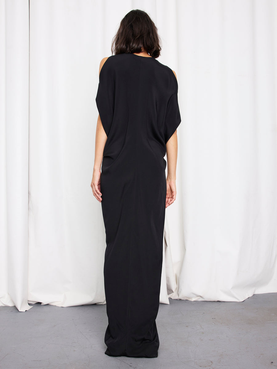 A person with short dark hair faces away, wearing the Zero + Maria Cornejo Long Miu Dress, a silk full-length style with cut-out shoulders. The background has white curtains and a gray floor. Made in New York.