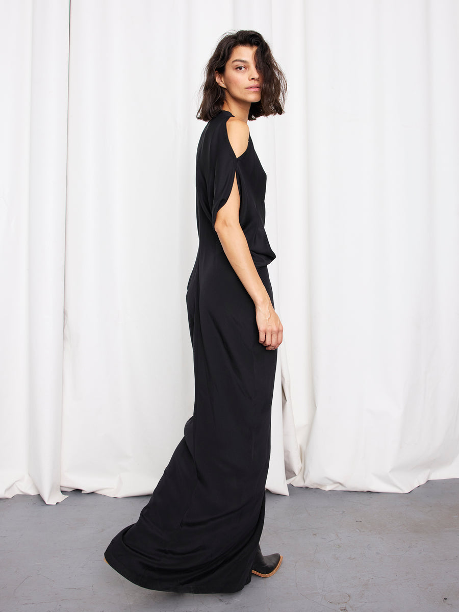 A woman with shoulder-length dark hair wears the Long Miu Dress by Zero + Maria Cornejo, a silk full-length design with cut-out shoulders, standing sideways before white curtains on a grey floor, looking at the camera. Made in New York.