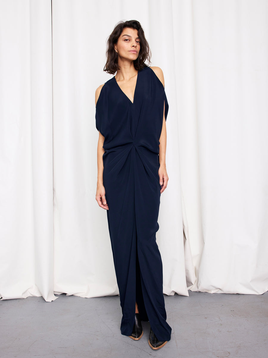 A woman with dark, shoulder-length hair and a neutral expression stands before white curtains, wearing the Zero + Maria Cornejo Long Miu Dress in navy silk with cut-out shoulders, deep V-neck, front knot, and pointed black shoes. Made in New York.