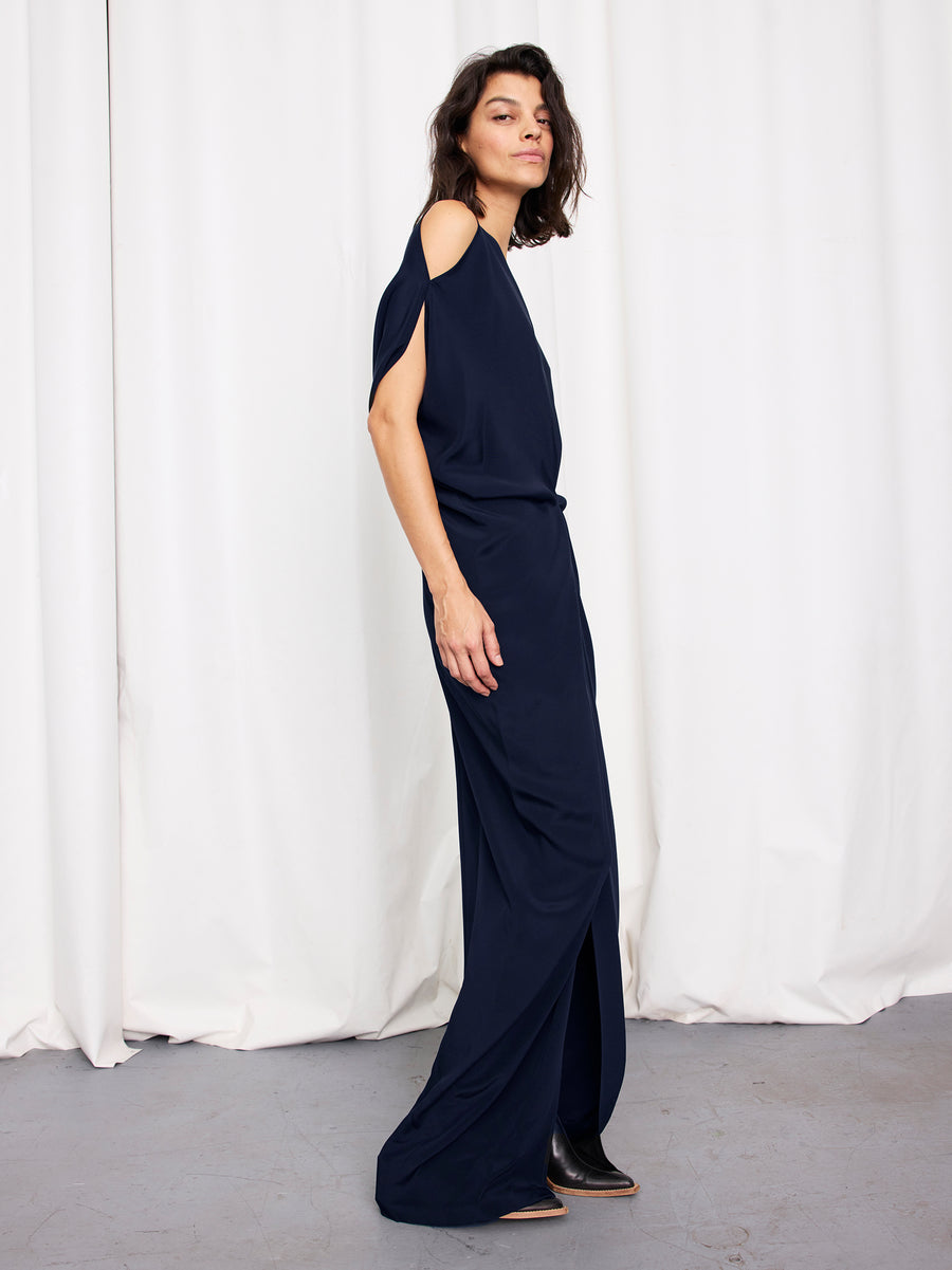 A person with medium-length dark hair stands sideways in the Zero + Maria Cornejo Long Miu Dress, a navy blue silk full-length dress with cut-out shoulders. Gray flooring and white curtains form the backdrop. Made in New York.