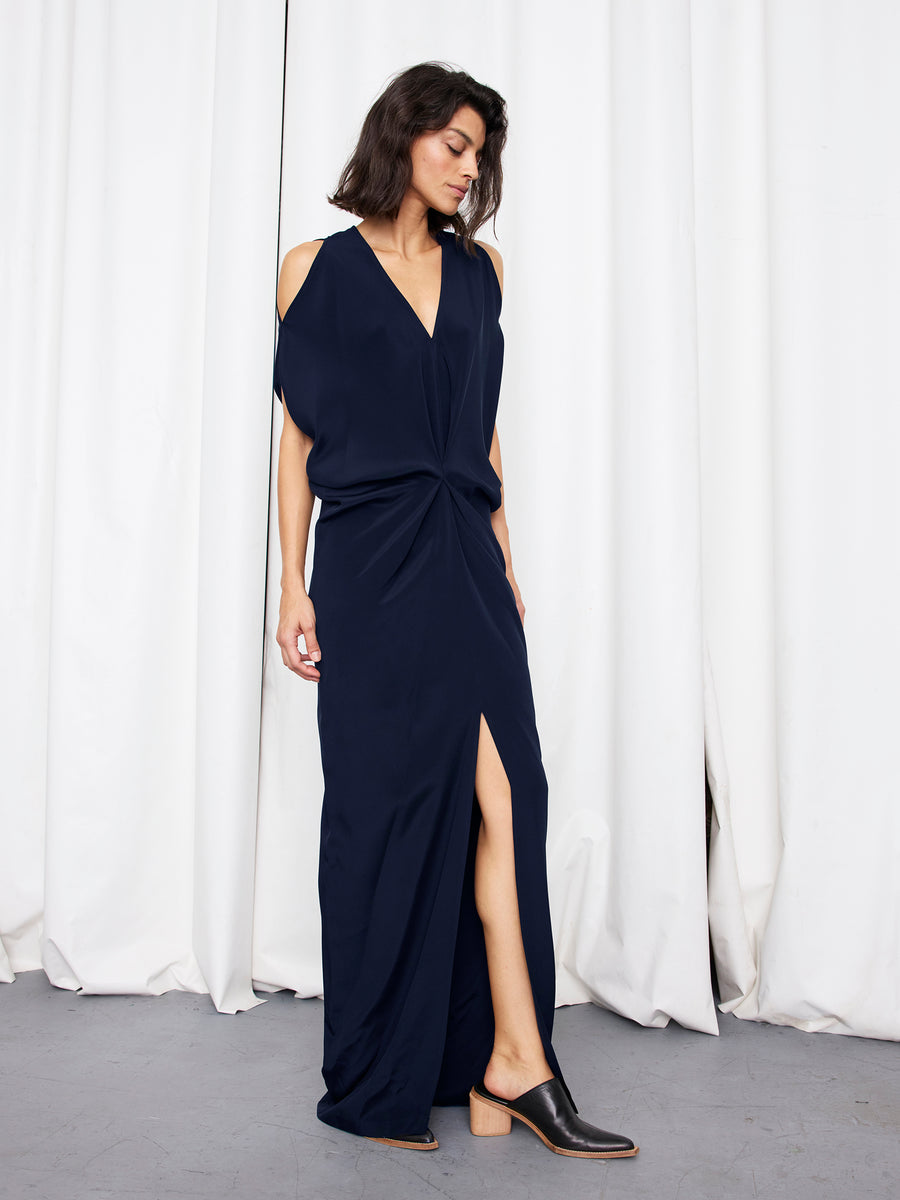 A person stands before white curtains wearing the Zero + Maria Cornejo Long Miu Dress in navy silk, featuring cut-out shoulders, V-neck, twisted waist, and a high front slit, paired with black slip-on shoes. Made in New York.