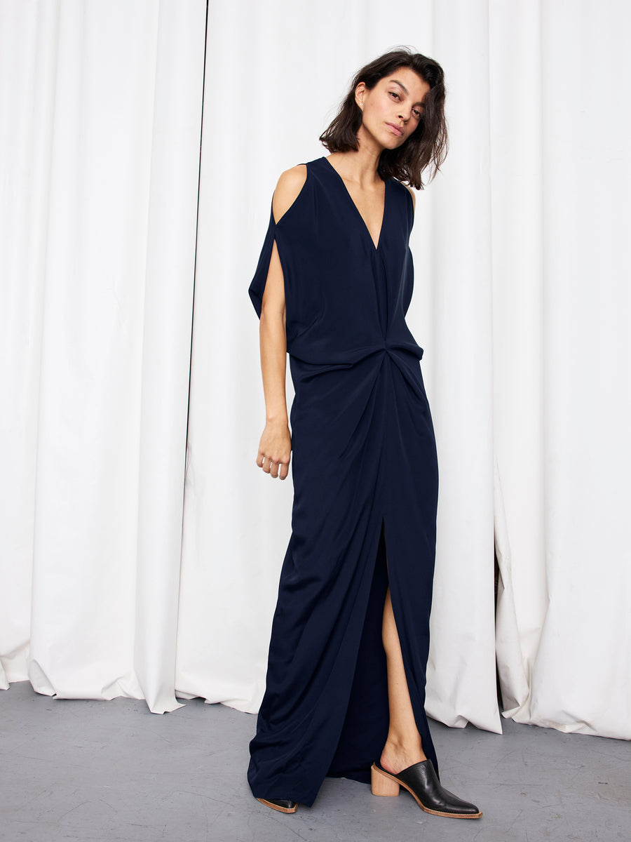 A woman models the Long Miu Dress by Zero + Maria Cornejo—navy blue silk, full-length with cut-out shoulders, front knot, and slit—standing before white curtains in black heeled mules. Made in New York.
