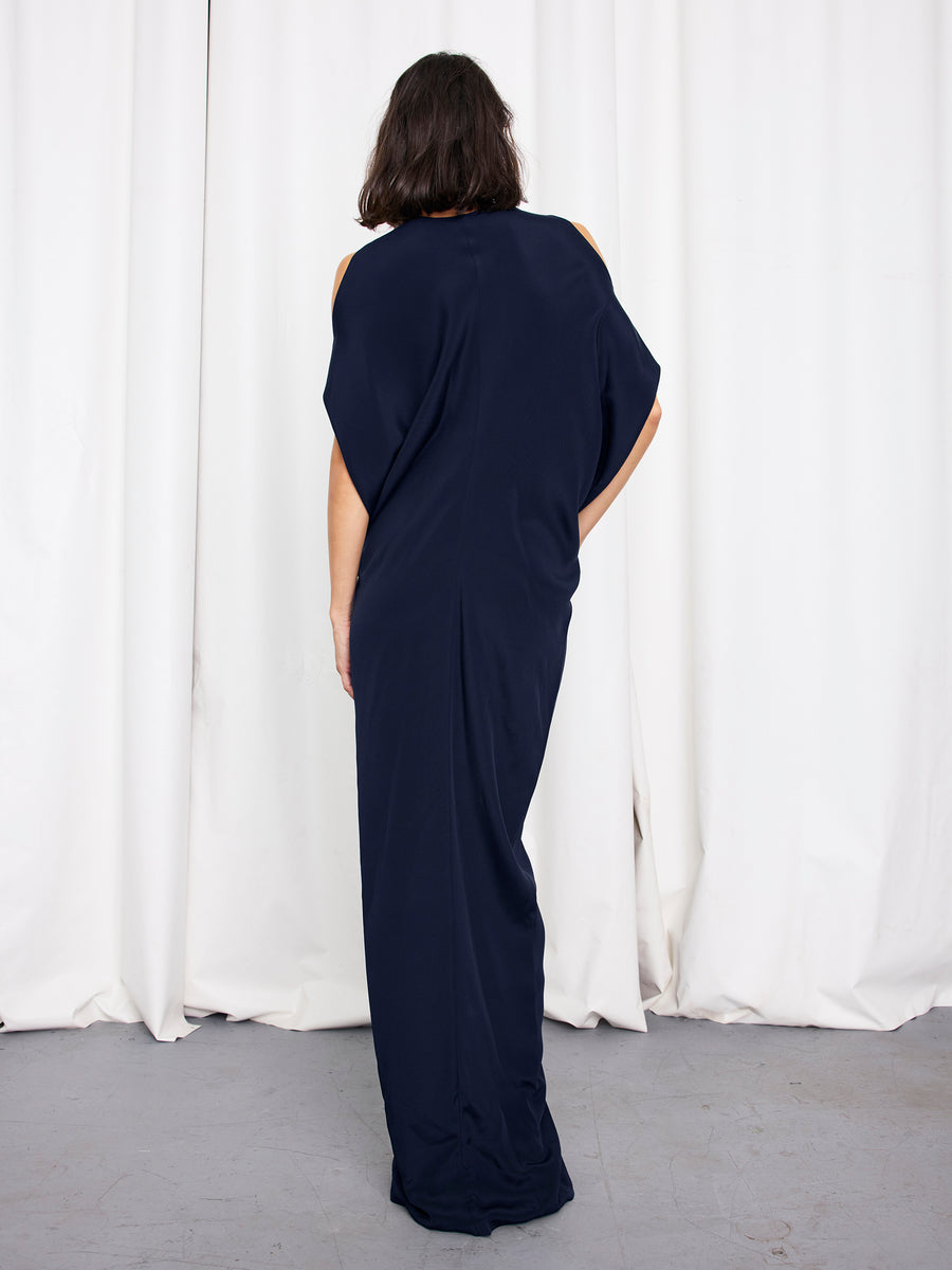 A person with shoulder-length dark hair, seen from behind, wears the Zero + Maria Cornejo Long Miu Dress—a silk, full-length style with cut-out shoulders. White curtains and a light gray floor complete the scene. Made in New York.