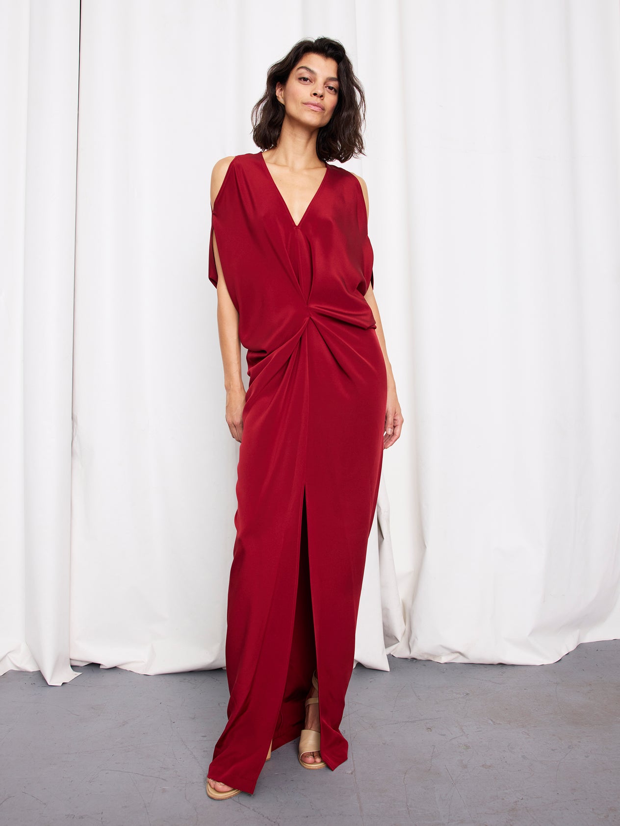 A woman walks confidently in front of white curtains wearing the Long Miu Dress by Zero + Maria Cornejo—a black, sleeveless, silk full-length dress with a deep V-neck and gathered knot detail that exudes sophistication and grace.