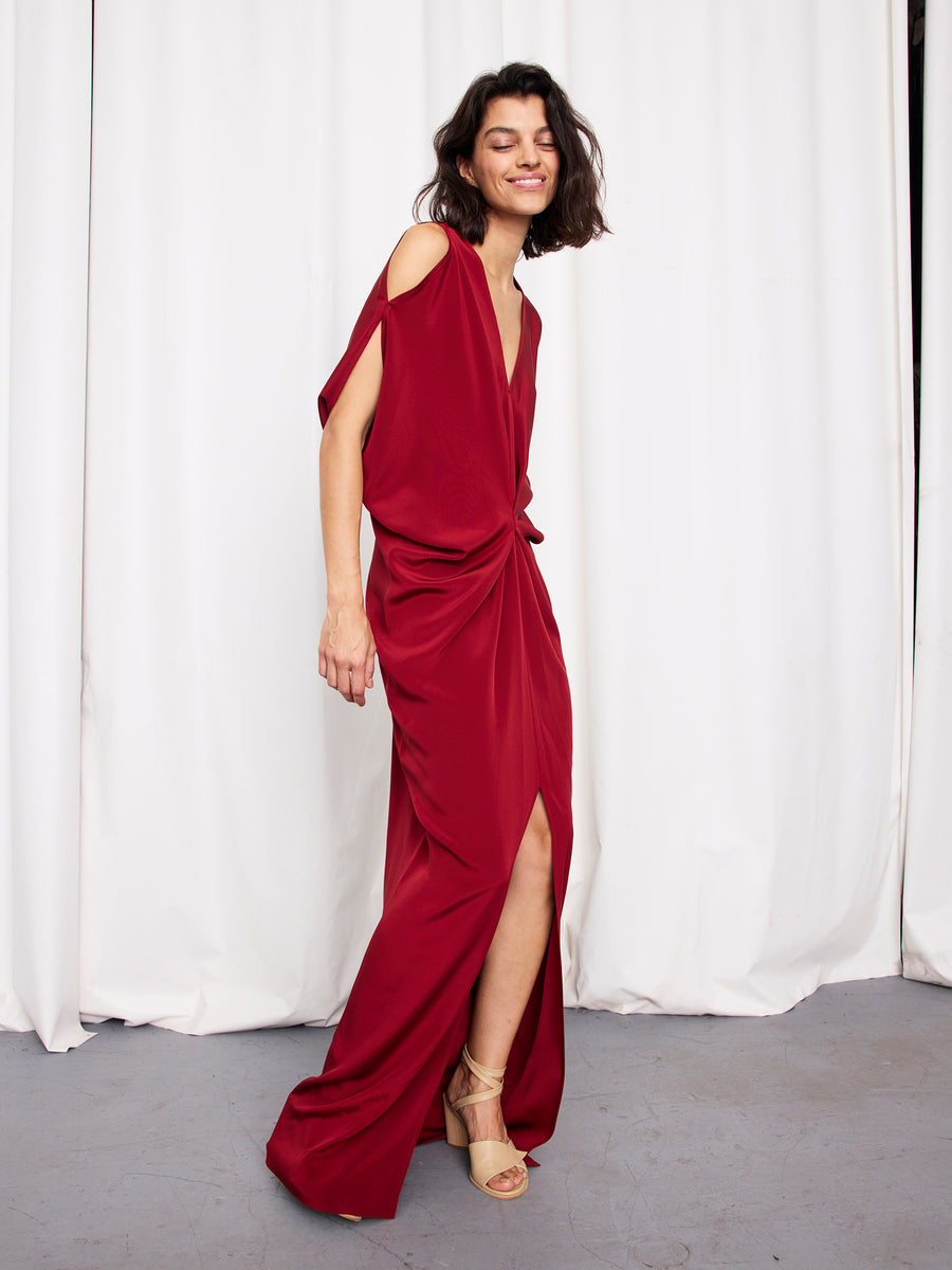 A woman in the Long Miu Dress by Zero + Maria Cornejo—a silk full-length dress with cut-out shoulders and a front slit—smiles, posing with one leg forward in beige heels. This chic look is proudly Made in New York.