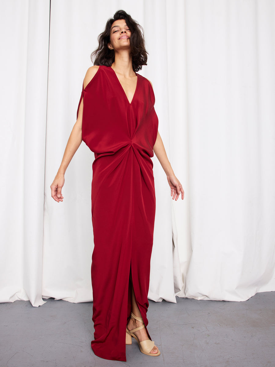 A woman in the Long Miu Dress by Zero + Maria Cornejo, a flowing deep red silk V-neck gown with a front knot, stands smiling before a white curtain. She has short dark hair and beige open-toe heels.