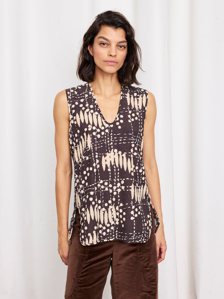 A woman with shoulder-length dark hair models the Ony Tank by Zero + Maria Cornejo, a sleeveless silk charmeuse top in black and beige patterns, paired with brown pants, against a white curtain backdrop.