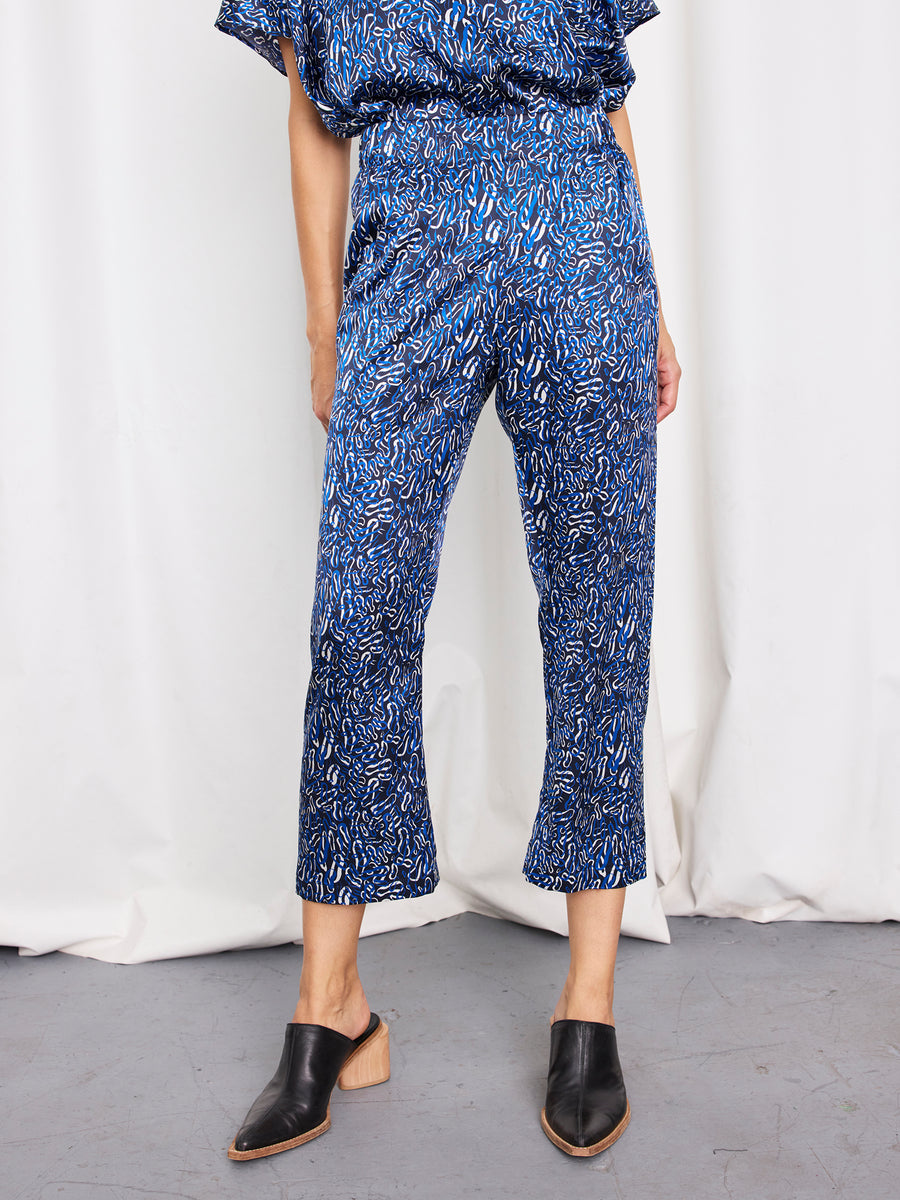 A person, visible from the shoulders down, stands in front of a white curtain wearing Zero + Maria Cornejo's Eko Pant—cropped blue patterned stretch silk charmeuse trousers with an elastic waist—and black heeled mules.
