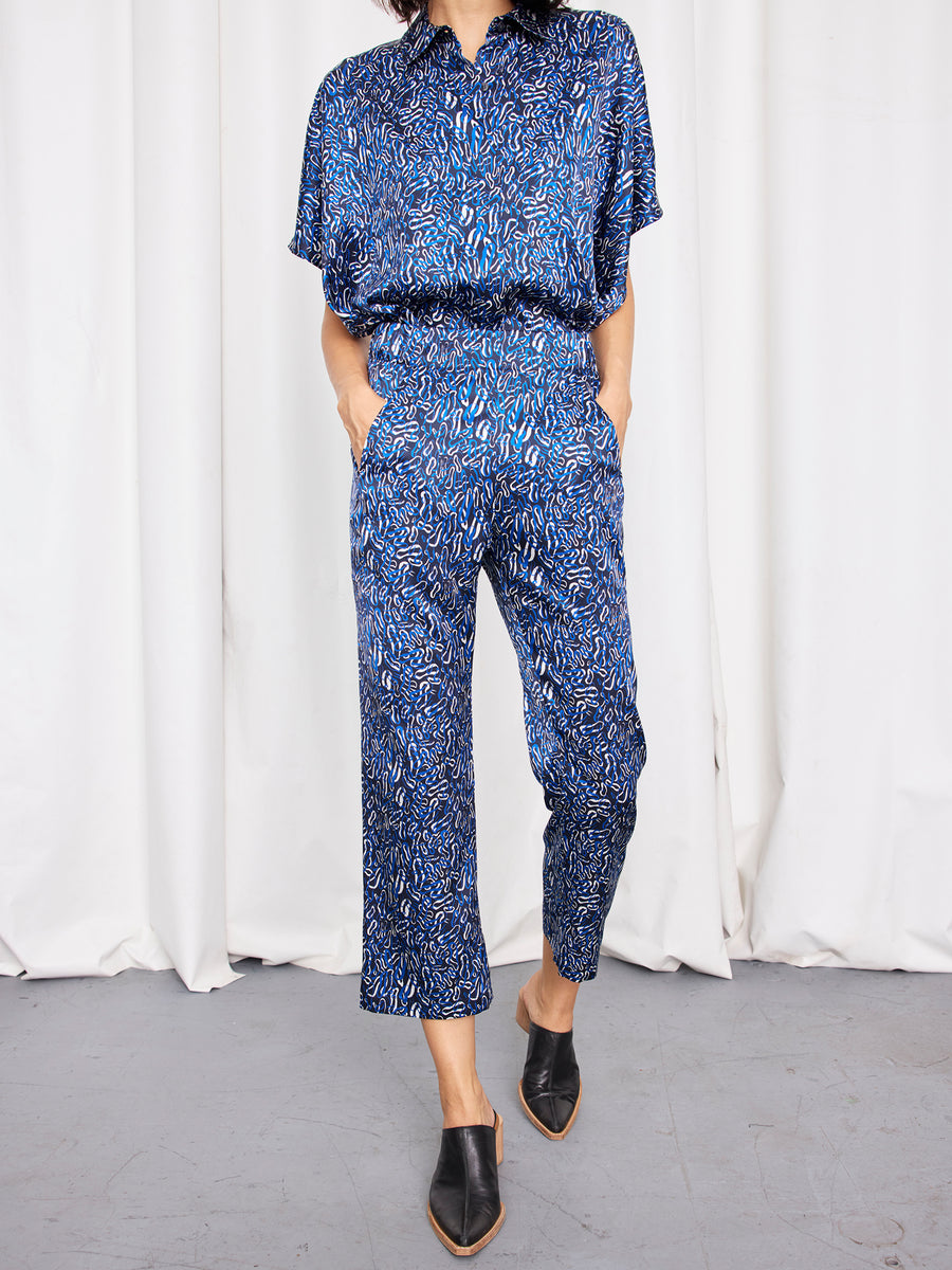 A person stands against white curtains wearing Zero + Maria Cornejo's Eko Pant—high-waisted stretch silk charmeuse trousers with an elastic waist—paired with a matching short-sleeve blue patterned blouse and black pointed-toe mules.
