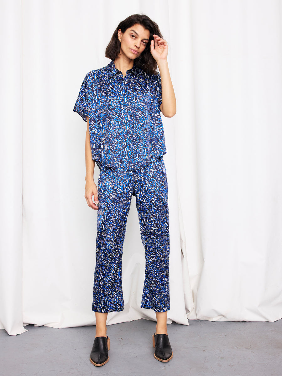A person stands before white curtains in a loose-fitting Zero + Maria Cornejo Circle Shirt with matching cropped pants and black slip-on shoes, right hand touching their hair.