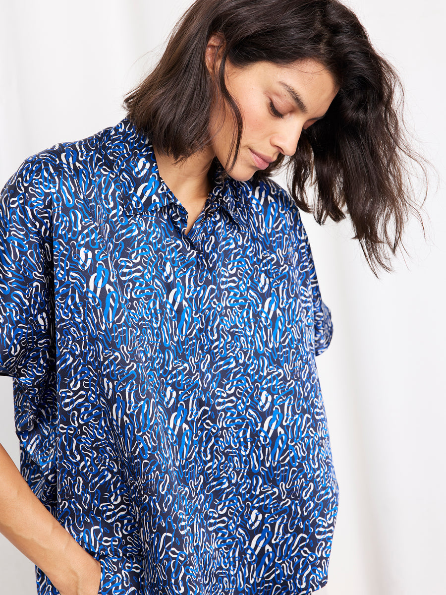 A woman with shoulder-length dark hair looks down, hands in her pockets, wearing the Circle Shirt by Zero + Maria Cornejo—a loose-fitting printed silk top—against a plain white background.