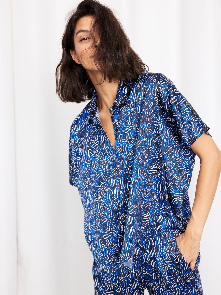A woman with shoulder-length dark hair models the Circle Shirt by Zero + Maria Cornejo—a loose, short-sleeved silk top with an abstract white and blue print—made in New York, posed against a white background.