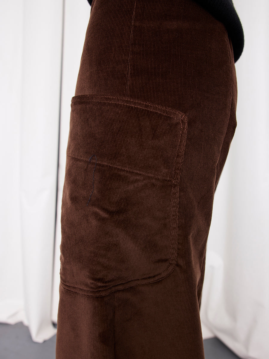 Close-up of a person wearing the Zero + Maria Cornejo Curved Eda Culotte, a stretch corduroy cropped pant made in New York with a large side patch pocket. Only the lower torso and upper leg are shown before white curtains.
