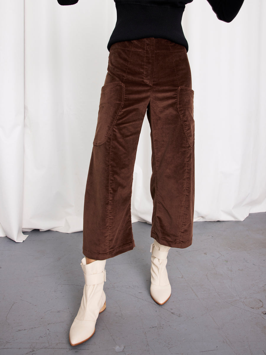 A person models the Zero + Maria Cornejo Curved Eda Culotte—brown stretch corduroy cropped pants made in New York—paired with a black sweater and white ankle boots, standing against a white curtain backdrop.