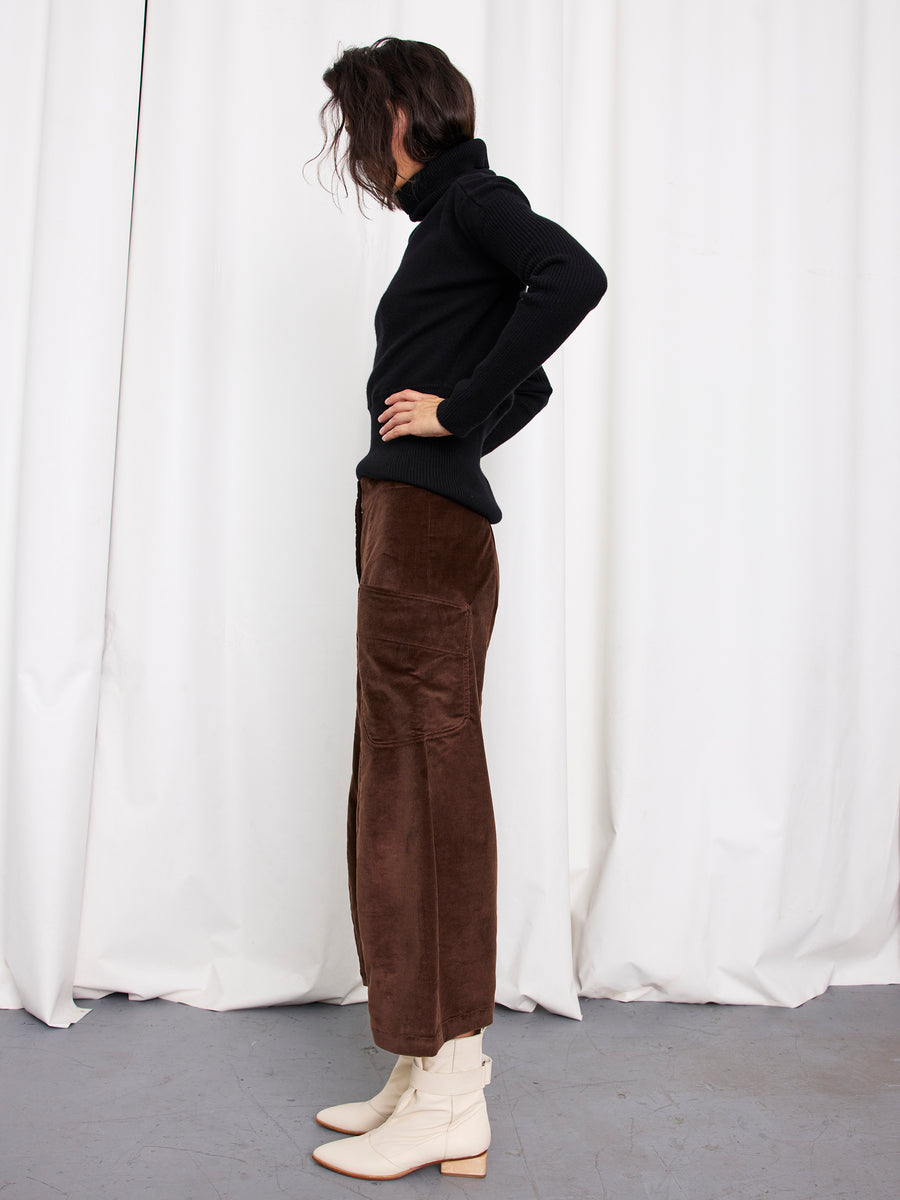 A person stands in profile against white curtains, wearing a black turtleneck and Zero + Maria Cornejo’s Curved Eda Culotte, styled with off-white ankle boots. Their hands rest on their hips while wavy, chin-length hair frames the look.