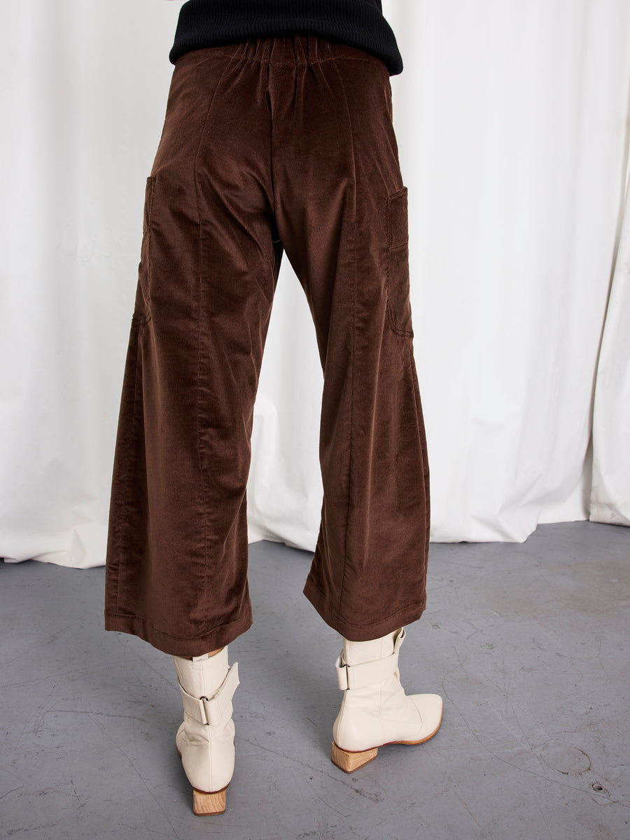 A person stands indoors, photographed from behind with a white curtain, wearing Zero + Maria Cornejo Curved Eda Culotte in brown corduroy and off-white ankle boots. The pants are cropped, stretch fabric, and made in New York.