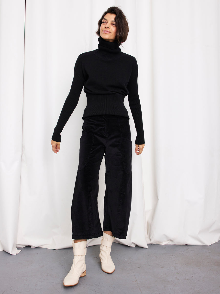 A person with short dark hair stands before white curtains, wearing a black turtleneck sweater and the Curved Eda Culotte by Zero + Maria Cornejo, paired with off-white ankle boots and exuding a relaxed, confident posture.