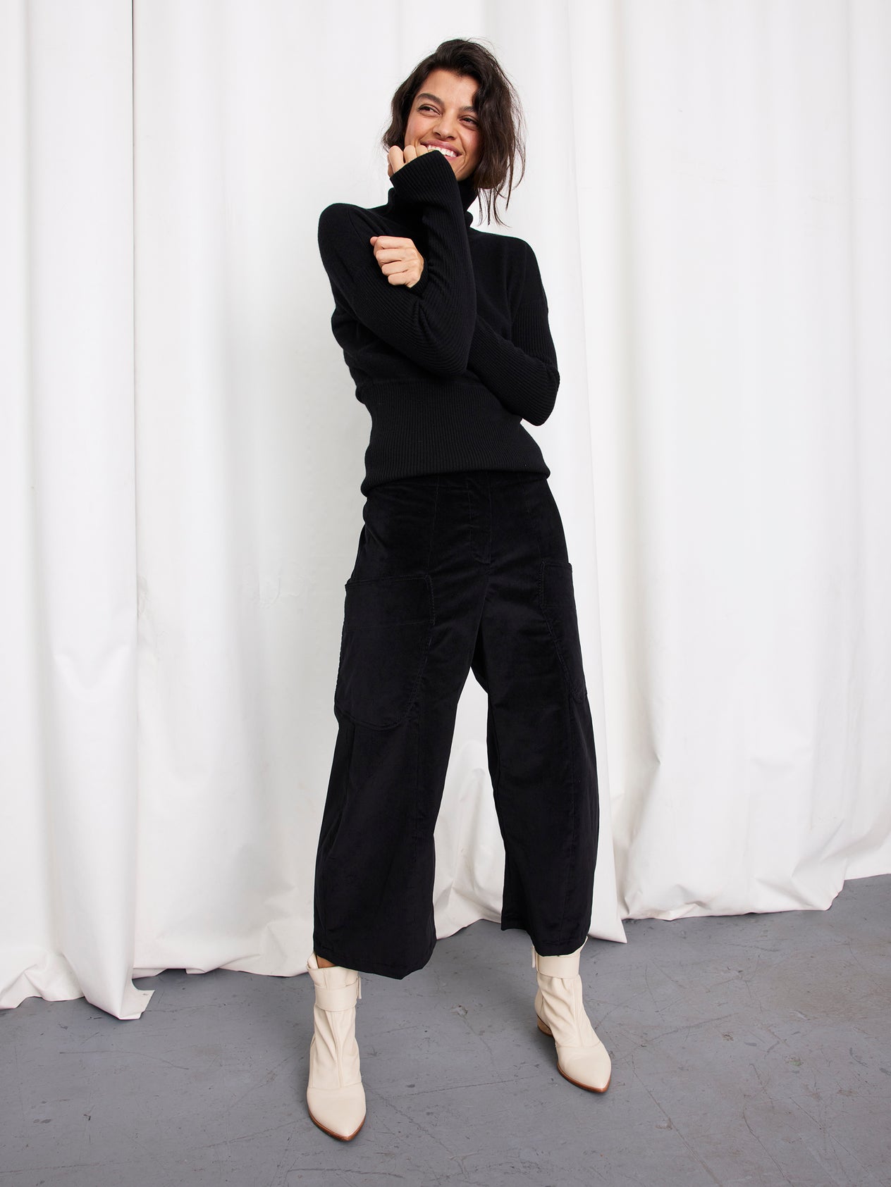 A person with dark hair, wearing a black long-sleeve top, Zero + Maria Cornejo’s Curved Eda Culotte made in New York, and white ankle boots, smiles in front of white curtains on a gray floor.