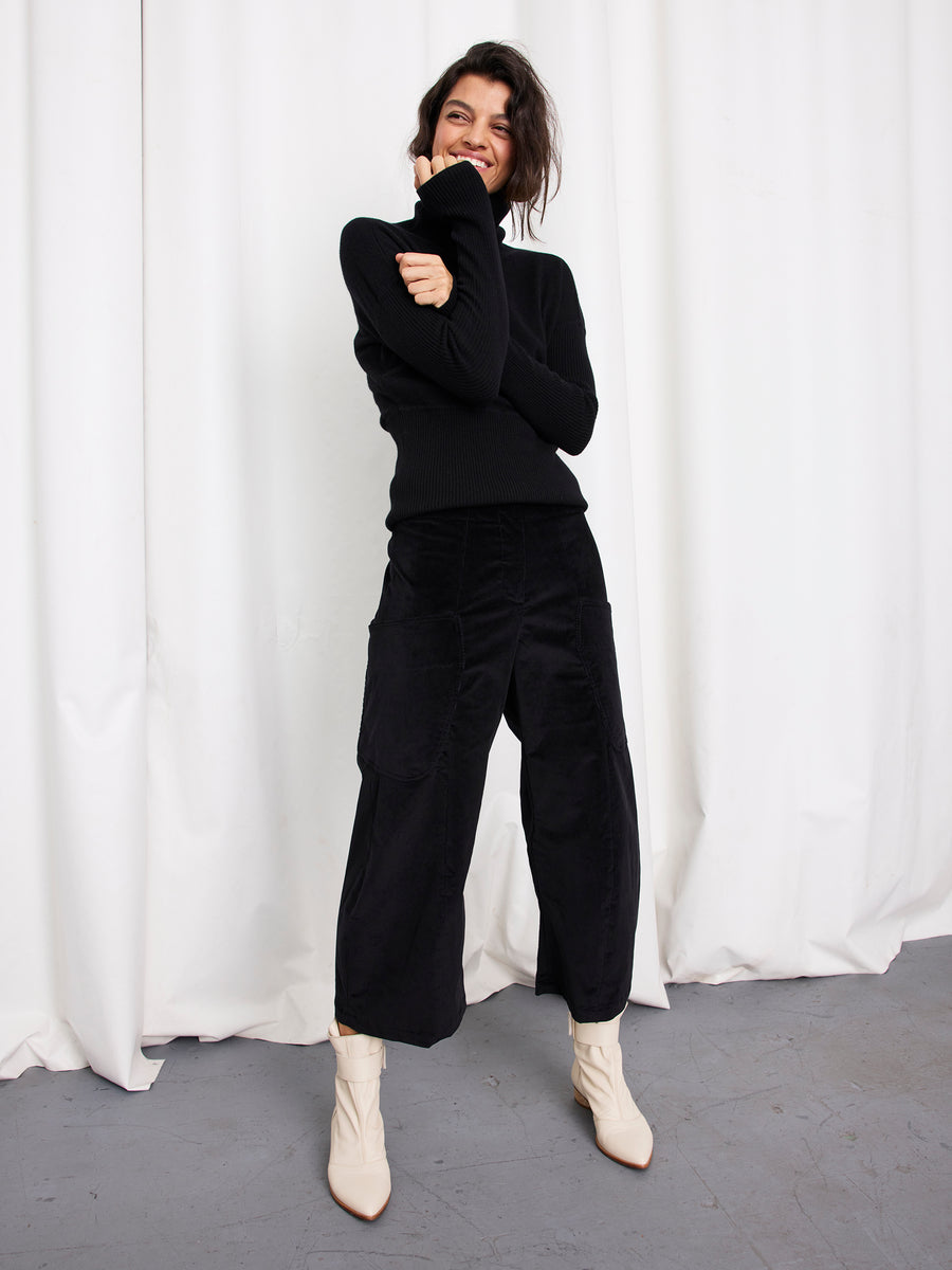 A person with dark hair, wearing a black long-sleeve top, Zero + Maria Cornejo’s Curved Eda Culotte made in New York, and white ankle boots, smiles in front of white curtains on a gray floor.