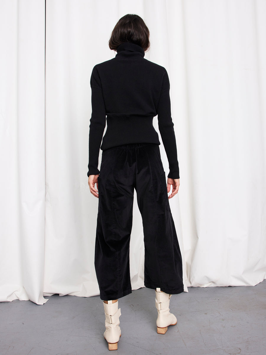 A person with short dark hair, wearing a black turtleneck and the Curved Eda Culotte by Zero + Maria Cornejo, stands facing away in front of white curtains on a gray floor.