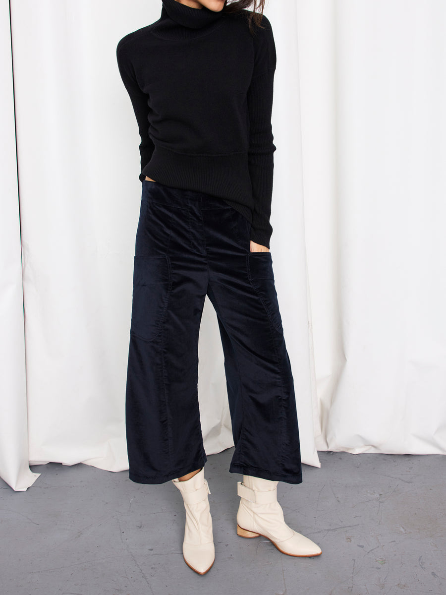 A person, face not visible, stands on a gray floor before white curtains wearing a black turtleneck, white ankle boots, and Zero + Maria Cornejo's Curved Eda Culotte stretch corduroy pants made in New York.