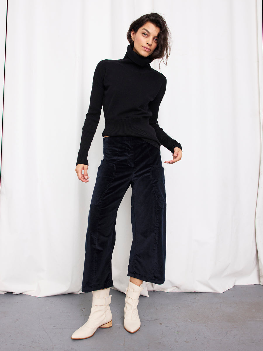 A person stands in front of white curtains wearing a black turtleneck, the Curved Eda Culotte by Zero + Maria Cornejo, and off-white ankle boots. They pose confidently with one hand on their hip.