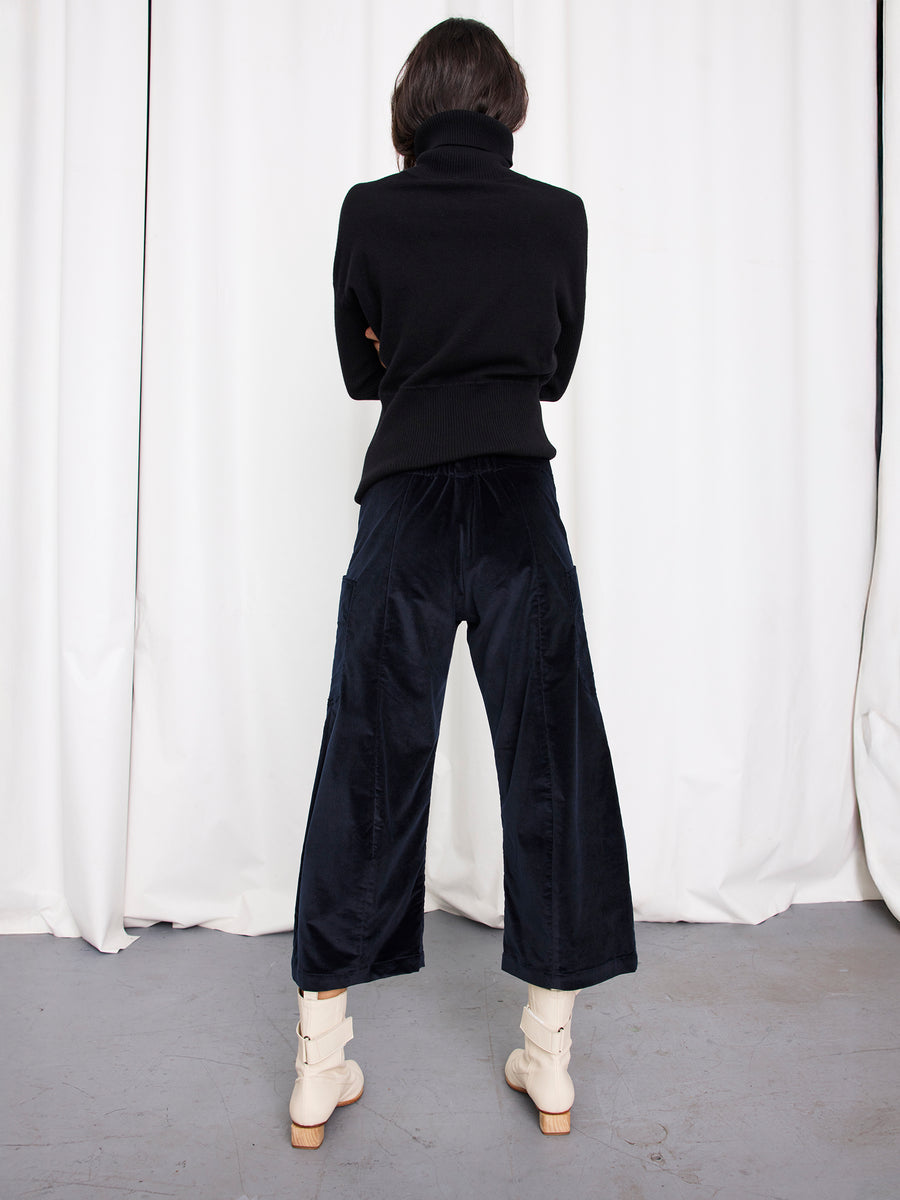 A person stands facing away, wearing a black turtleneck, Curved Eda Culotte by Zero + Maria Cornejo, and cream ankle boots in front of white curtains on a gray floor.