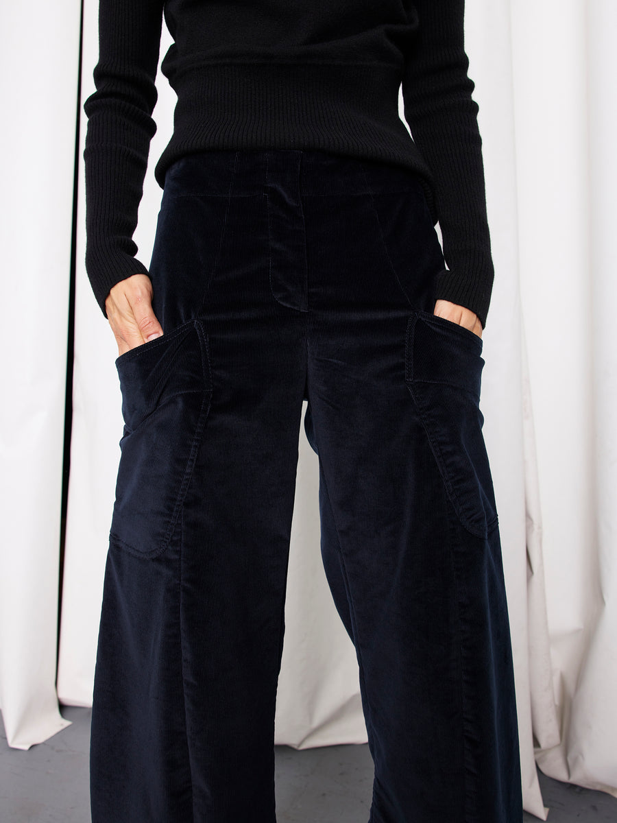 A person wears a black long-sleeve top and the Zero + Maria Cornejo Curved Eda Culotte, featuring large front pockets, standing with hands in pockets before white curtains.