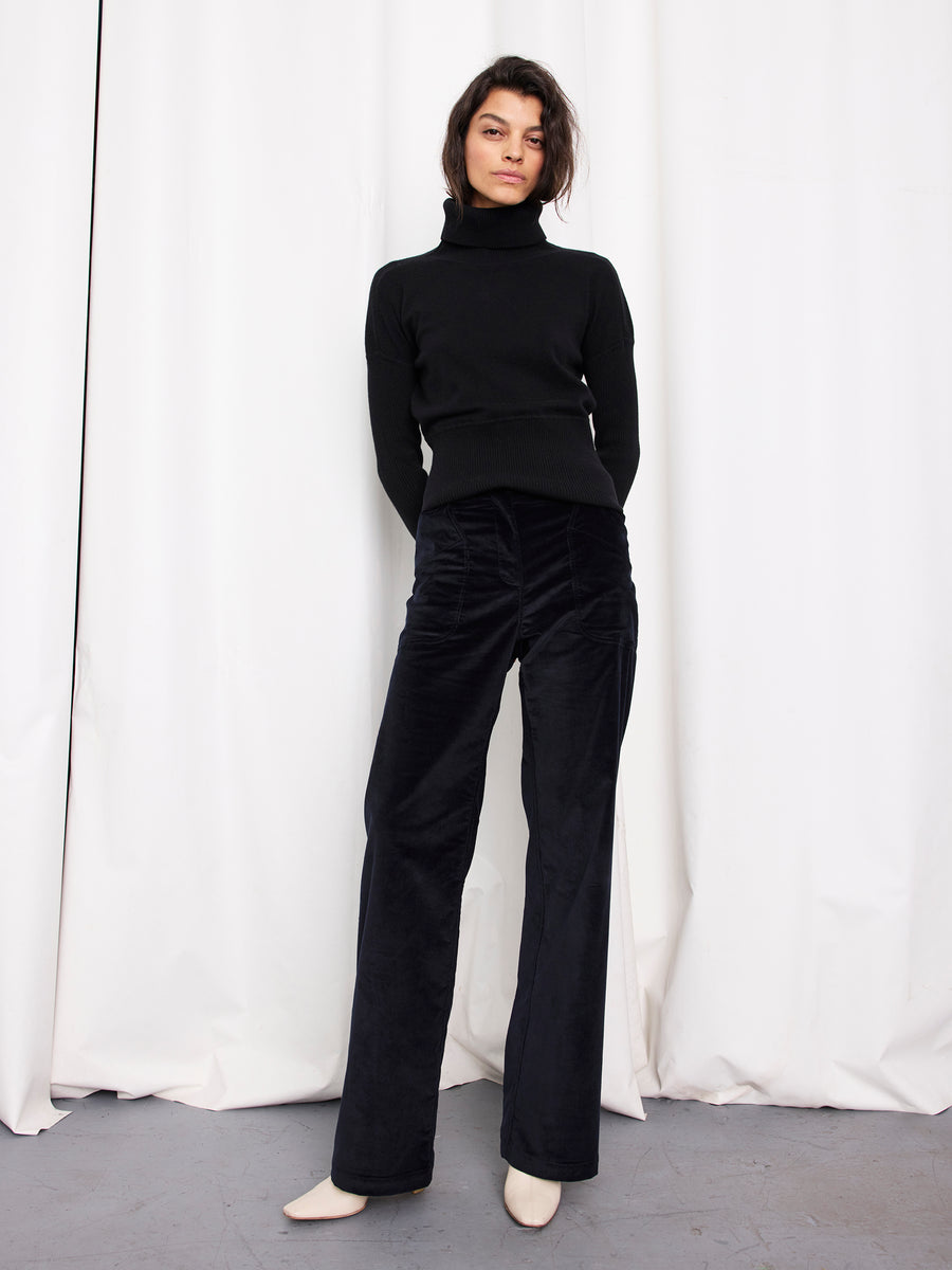 A person with short dark hair and a neutral expression stands before white curtains, wearing a black turtleneck, the Eda Pant by Zero + Maria Cornejo in wide-leg organic cotton corduroy, and cream shoes.
