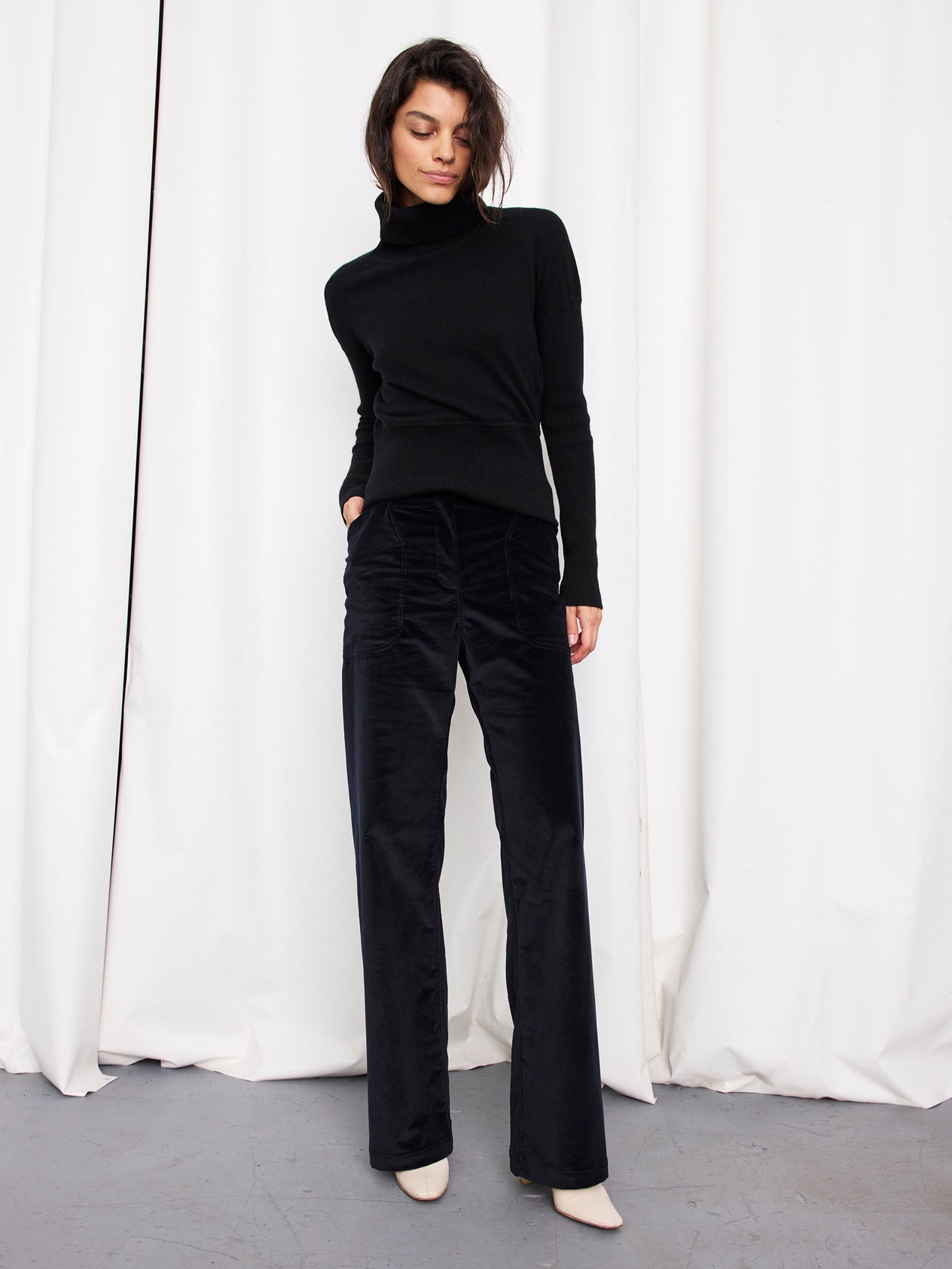 A woman stands in front of white curtains, wearing a black turtleneck sweater and Zero + Maria Cornejo Eda Pant in organic cotton wide-leg corduroy, with white shoes, looking down with one hand in her pocket.
