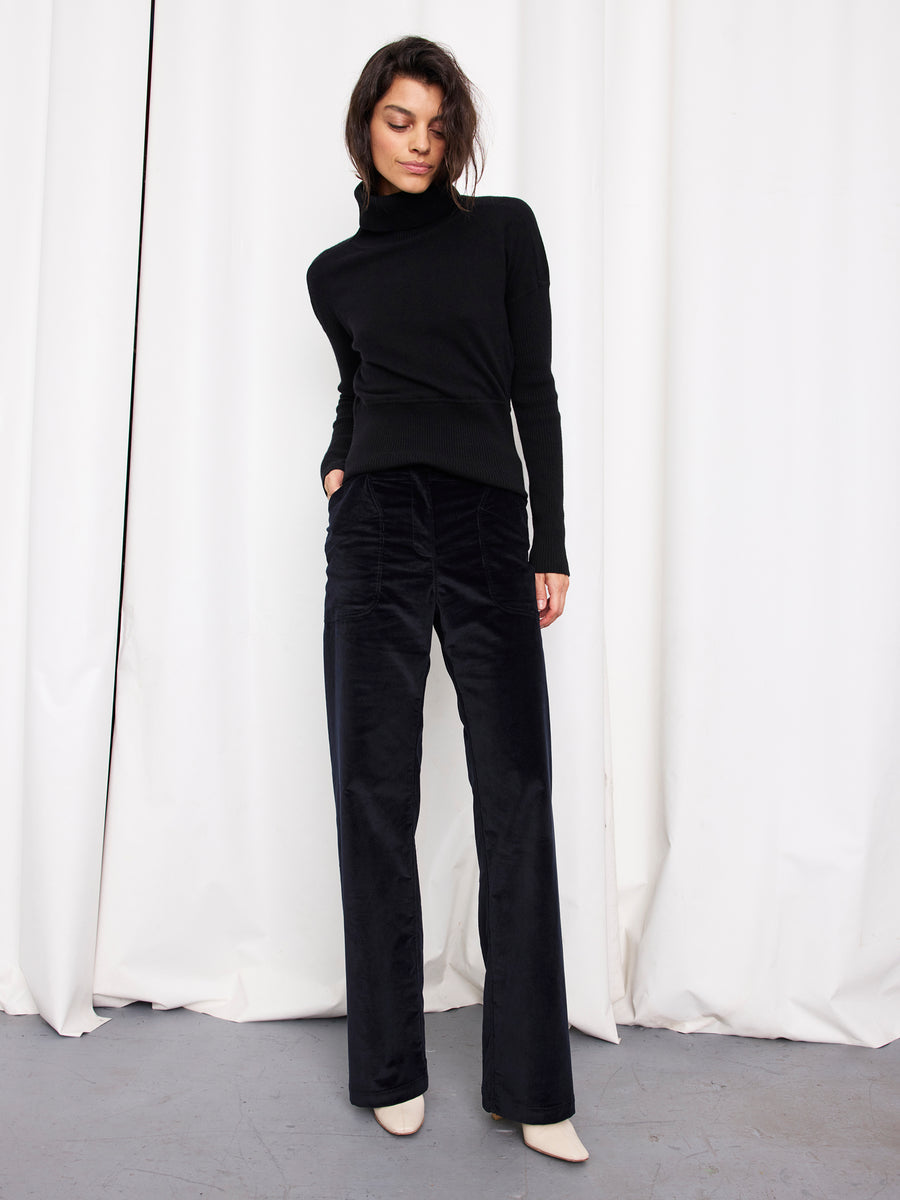 A woman stands in front of white curtains, wearing a black turtleneck sweater and Zero + Maria Cornejo Eda Pant in organic cotton wide-leg corduroy, with white shoes, looking down with one hand in her pocket.