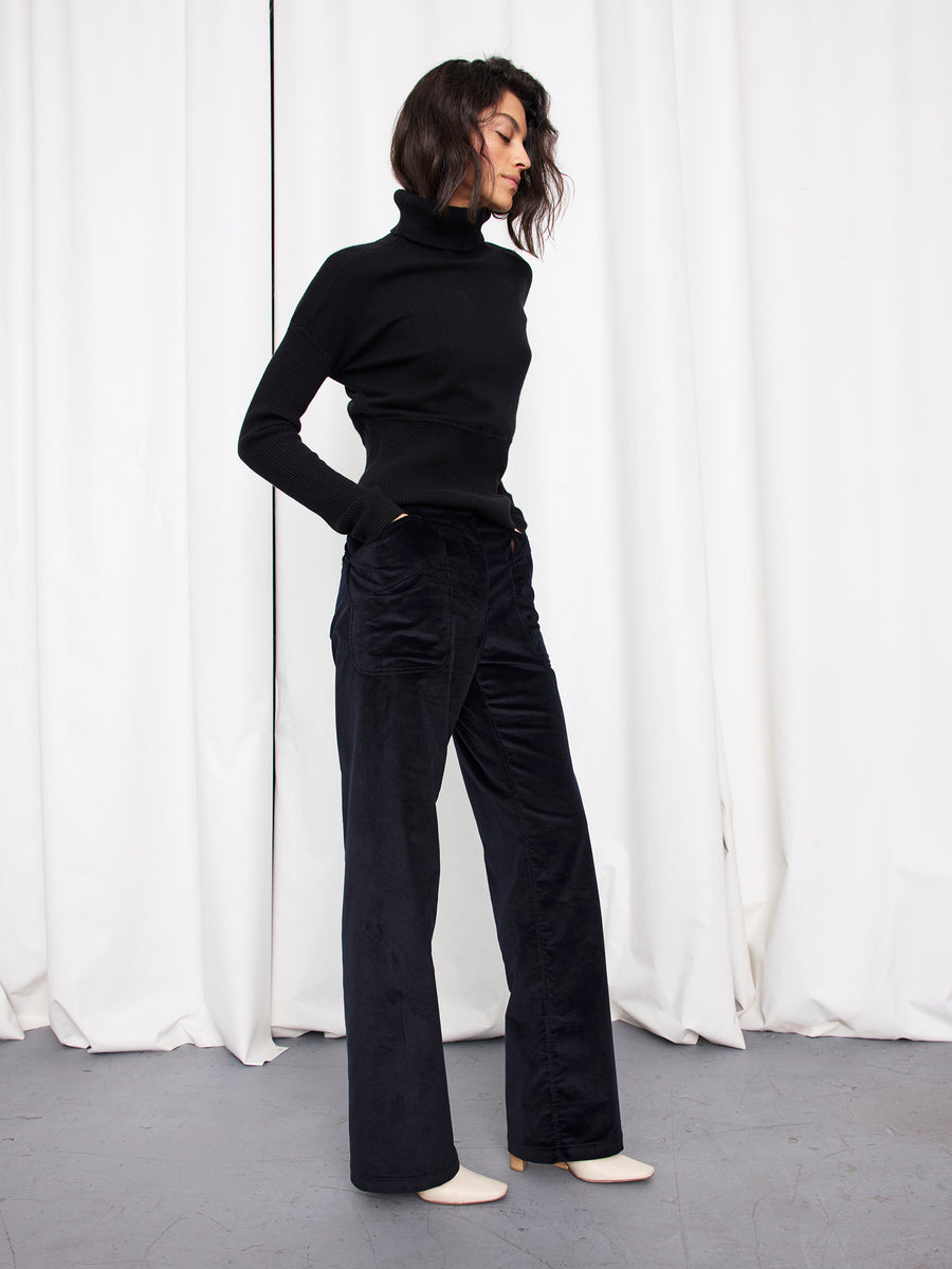 A woman stands in front of white curtains wearing a black turtleneck, the Zero + Maria Cornejo Eda Pant in organic cotton corduroy, and white shoes, with her hands in her pockets and looking to the side.