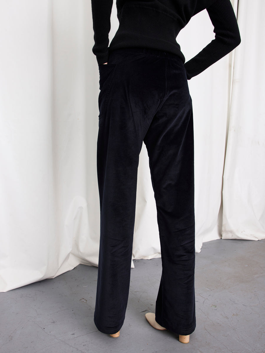 A person stands with their back to the camera, wearing the Eda Pant by Zero + Maria Cornejo—a black wide-leg velvet style—with their hand in one pocket, posed in front of white curtains on a gray floor.