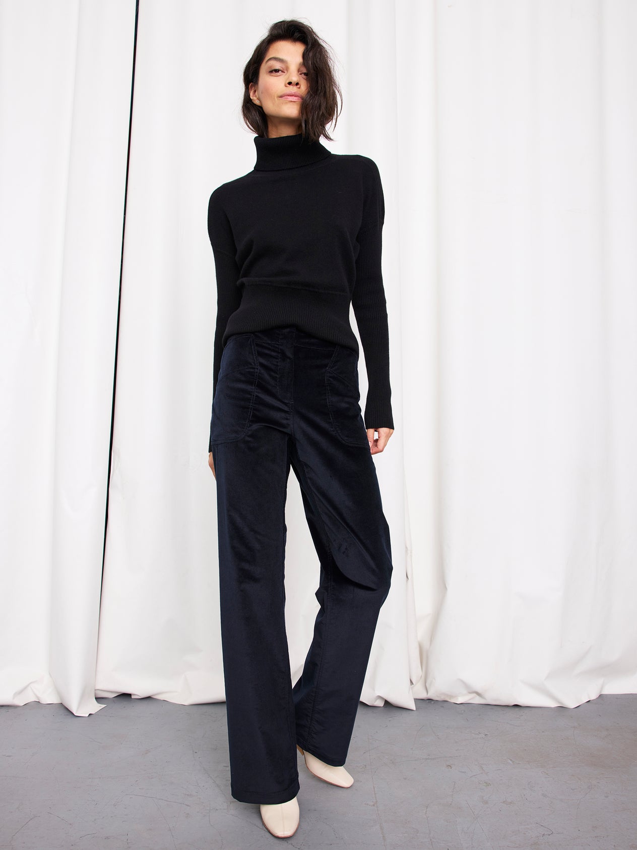 A woman stands in front of white curtains, wearing a black turtleneck sweater and Zero + Maria Cornejo Eda Pant in organic cotton wide-leg corduroy, with white shoes, looking down with one hand in her pocket.