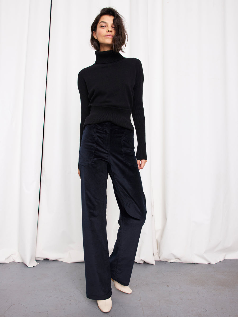A woman with short, dark hair stands confidently before white curtains, wearing a black turtleneck, Zero + Maria Cornejo Eda Pant in organic cotton, and cream shoes.