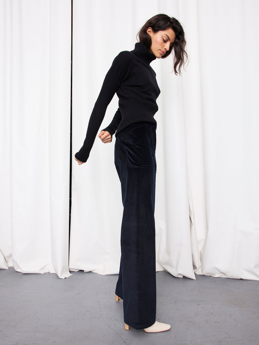 A woman stands in profile against white curtains, wearing the Eda Pant by Zero + Maria Cornejo with a black turtleneck sweater and white heeled shoes. She looks down, one arm bent and the other extended behind her.