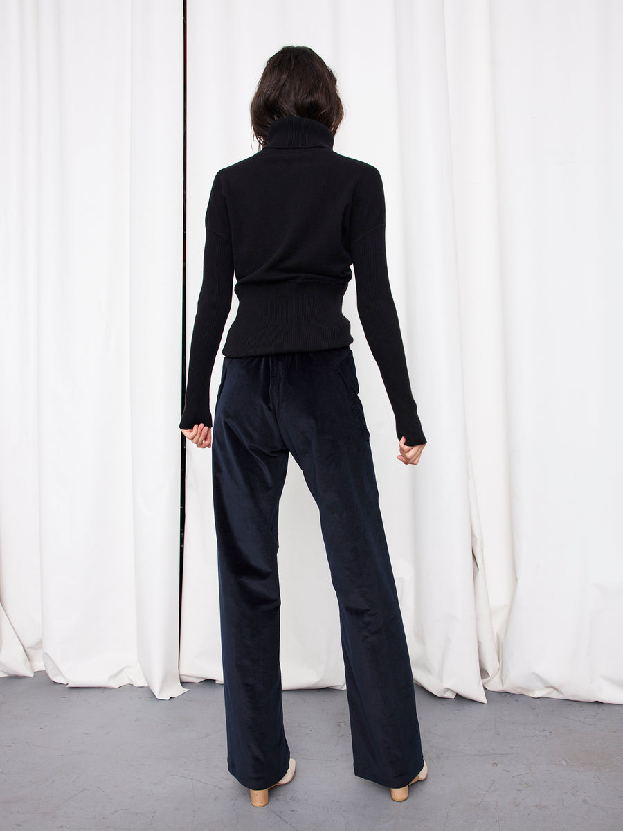 A person faces away from the camera, wearing a black turtleneck sweater and Zero + Maria Cornejo's Eda Pant—dark, wide-leg organic cotton corduroy—set against white curtains and a gray floor.