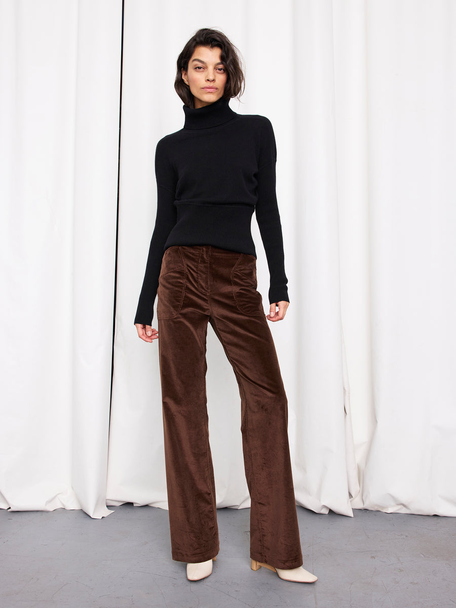 A woman stands against white curtains wearing a fitted black turtleneck, the brown Eda Pant by Zero + Maria Cornejo, and cream-colored heeled boots.