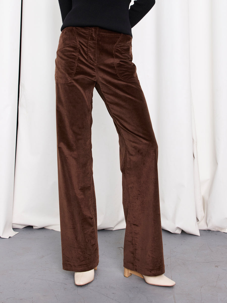 A person wears the Eda Pant by Zero + Maria Cornejo—high-waisted, wide-leg, brown corduroy made from organic cotton—with white heeled shoes, standing before white curtains on a gray floor; upper body mostly out of frame.