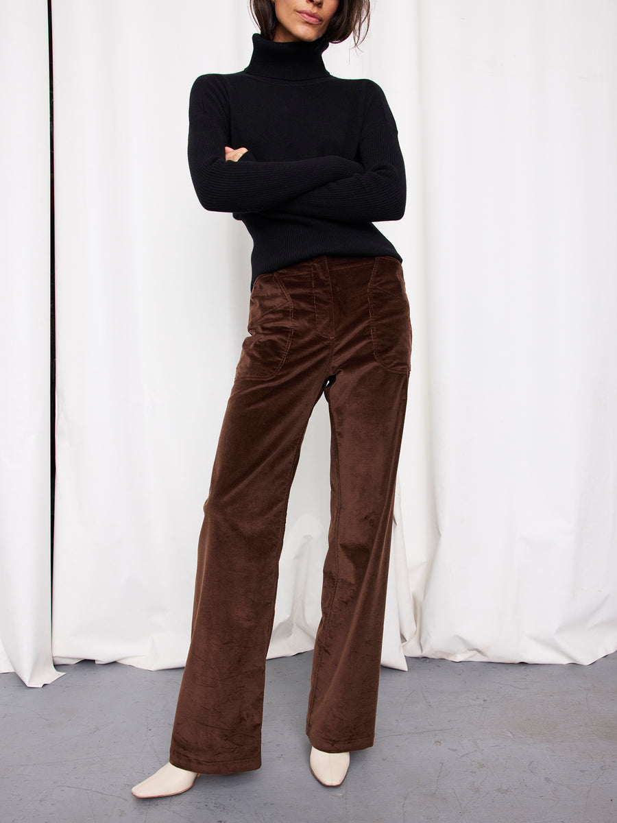 A person stands with arms crossed, wearing a black turtleneck sweater, Zero + Maria Cornejo's Eda Pant in high-waisted wide leg corduroy, and off-white shoes, posed in front of white curtains.