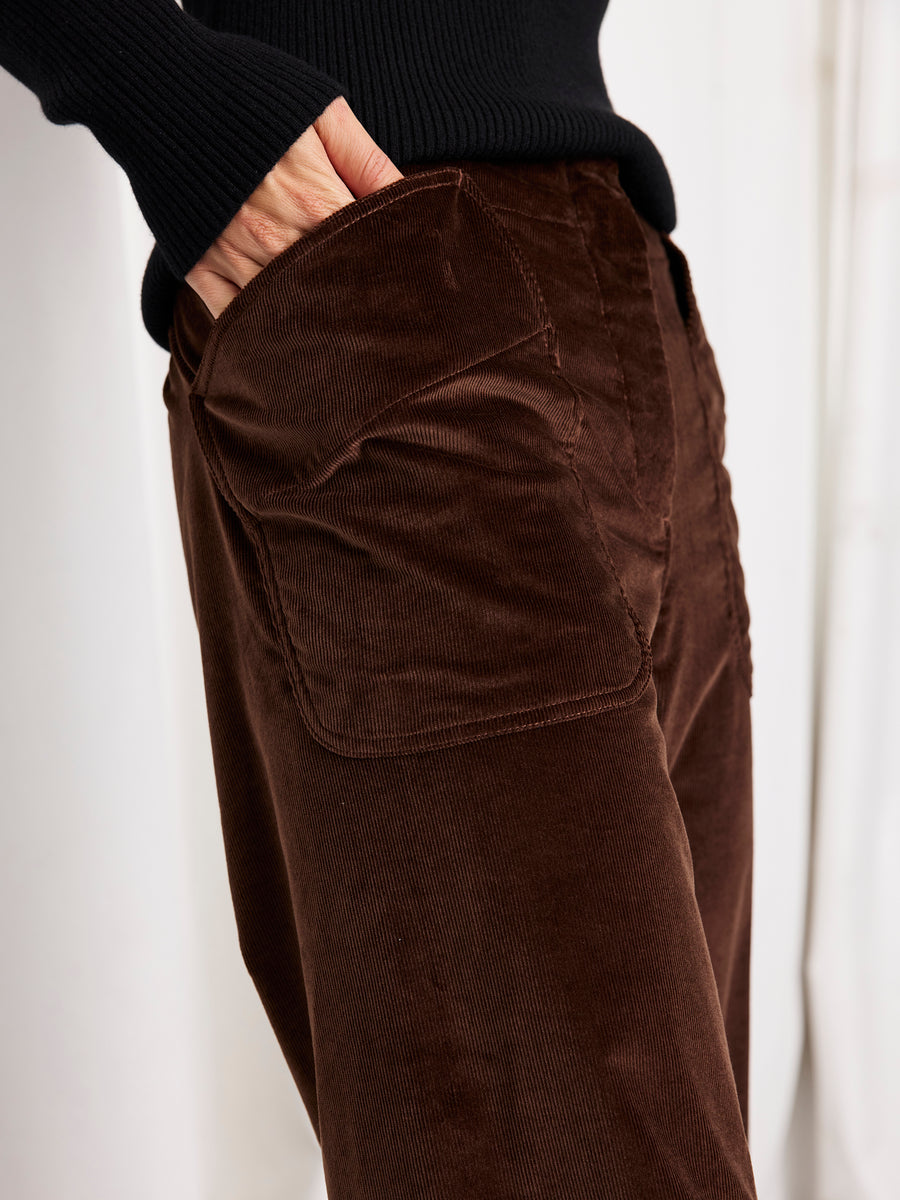 A person wearing the Eda Pant by Zero + Maria Cornejo—wide leg brown corduroy pants with large front pockets—and a black ribbed sweater, one hand tucked into a pocket.