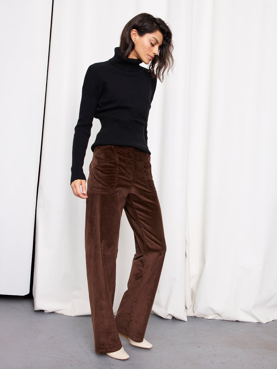 A woman stands before white curtains wearing the Zero + Maria Cornejo Eda Pant in brown velvet, a black turtleneck sweater, and cream-colored shoes, her head slightly bowed and hands at her sides.