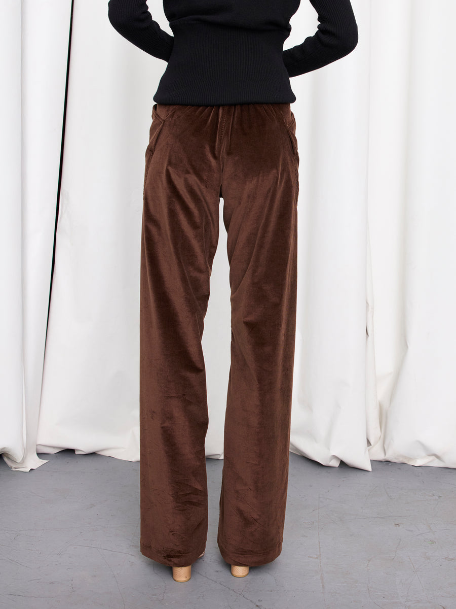 Only the lower half of a person is visible, showing them wearing the Eda Pant by Zero + Maria Cornejo—high-waisted, wide-leg brown corduroy pants—paired with a black long-sleeve top against white curtains.
