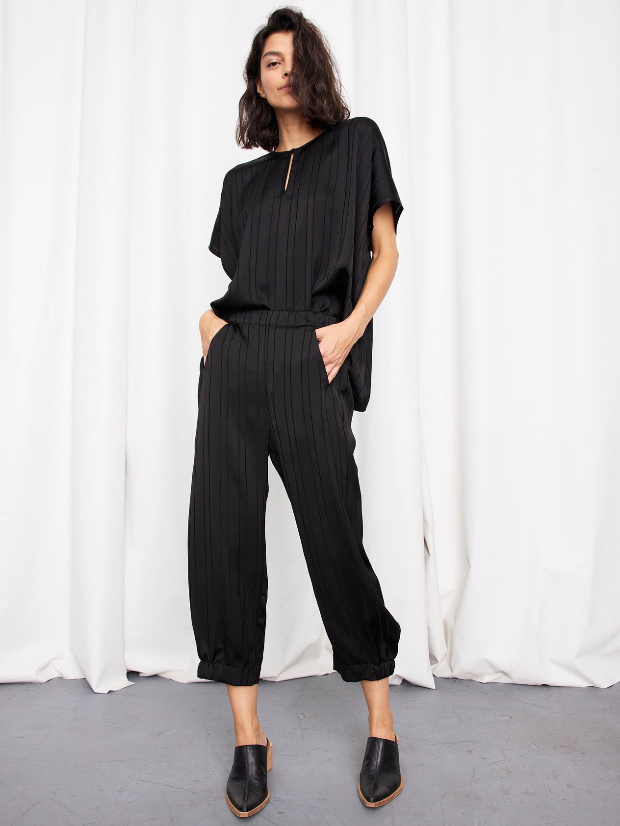A woman stands before white curtains wearing the Abi Pant by Zero + Maria Cornejo, a loose-fitting, woven-striped black cropped pant with pockets. She pairs it with black heeled mules. Made in New York.