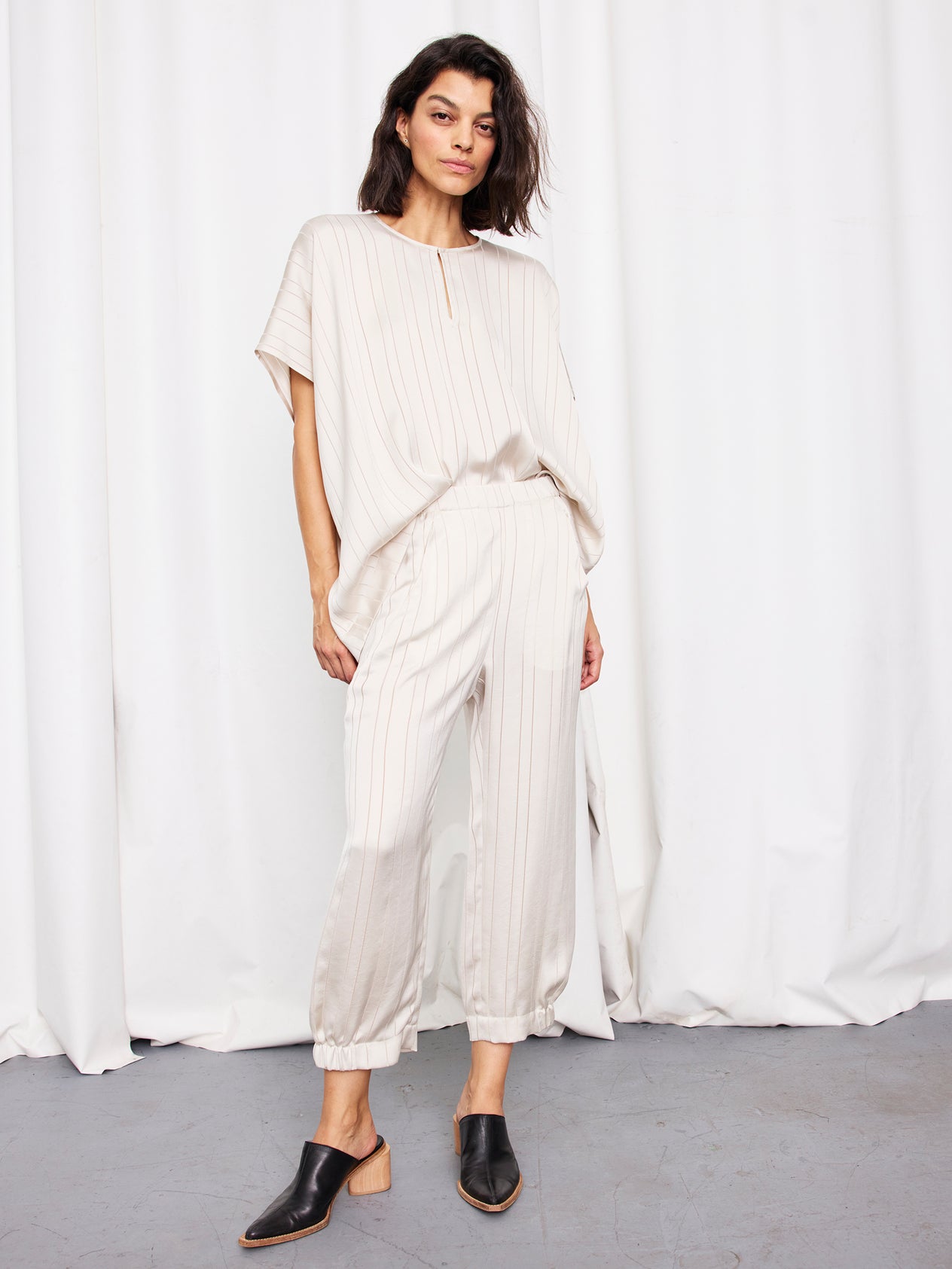 A woman stands before white curtains wearing the Abi Pant by Zero + Maria Cornejo, a loose-fitting, woven-striped black cropped pant with pockets. She pairs it with black heeled mules. Made in New York.