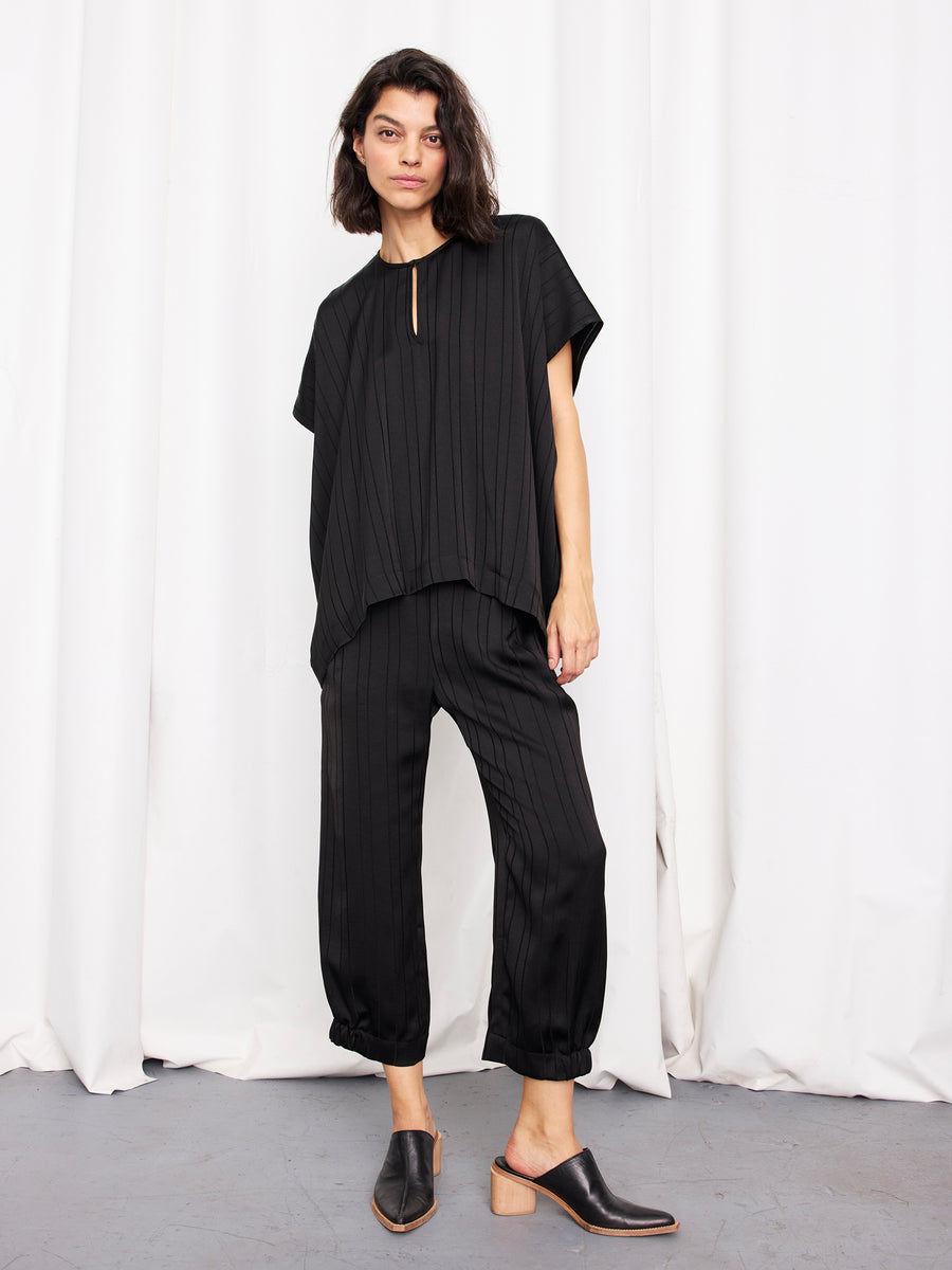 A person stands before white curtains wearing the Rae Poncho Top by Zero + Maria Cornejo, paired with black pinstripe pants and mule-style shoes. This elegant look is thoughtfully made in New York.