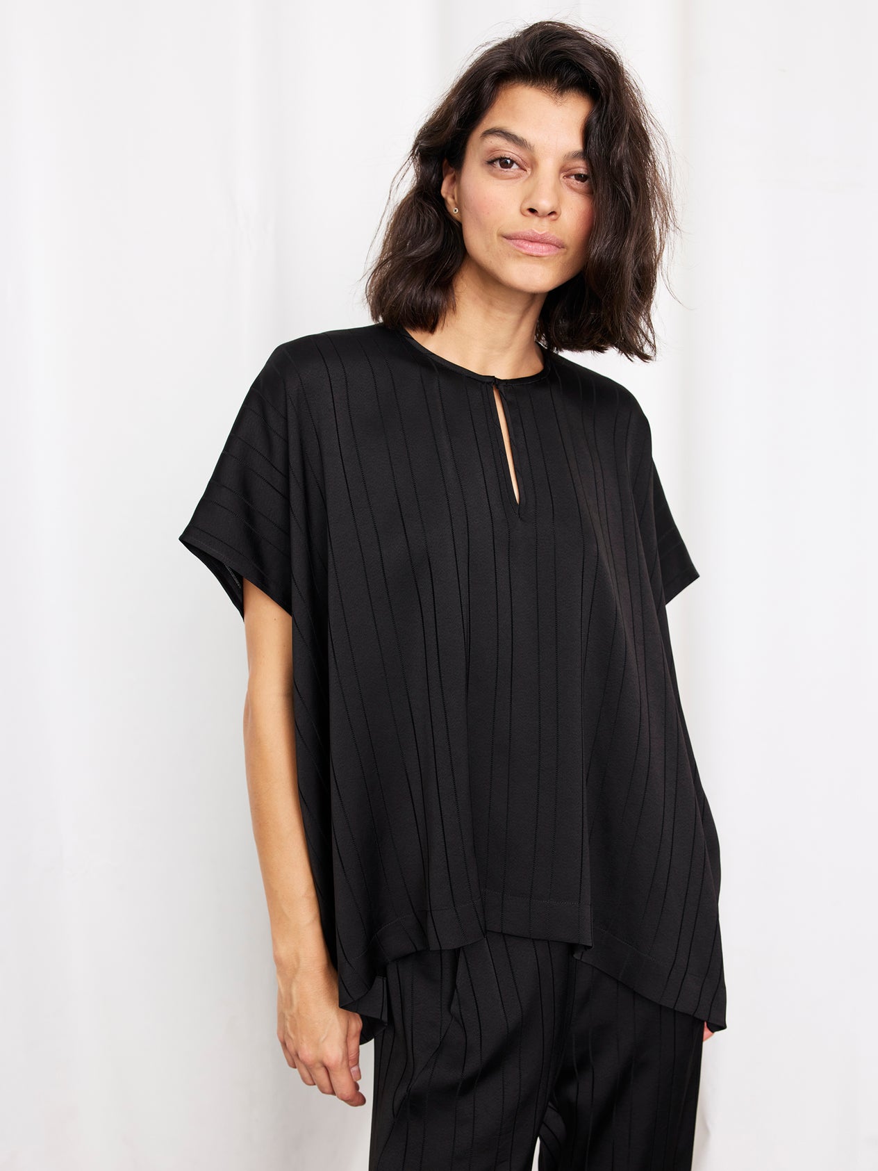 A woman with shoulder-length dark hair wears the Zero + Maria Cornejo Rae Poncho Top, a woven striped draped piece, paired with black pants and standing against a plain white background. This stylish look is made in New York.