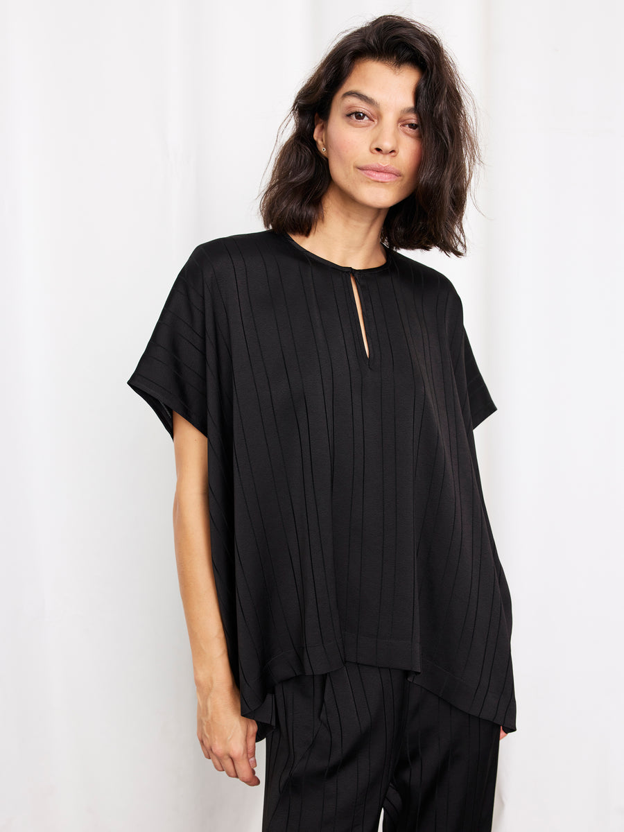 A woman with shoulder-length dark hair wears the Zero + Maria Cornejo Rae Poncho Top, a woven striped draped piece, paired with black pants and standing against a plain white background. This stylish look is made in New York.