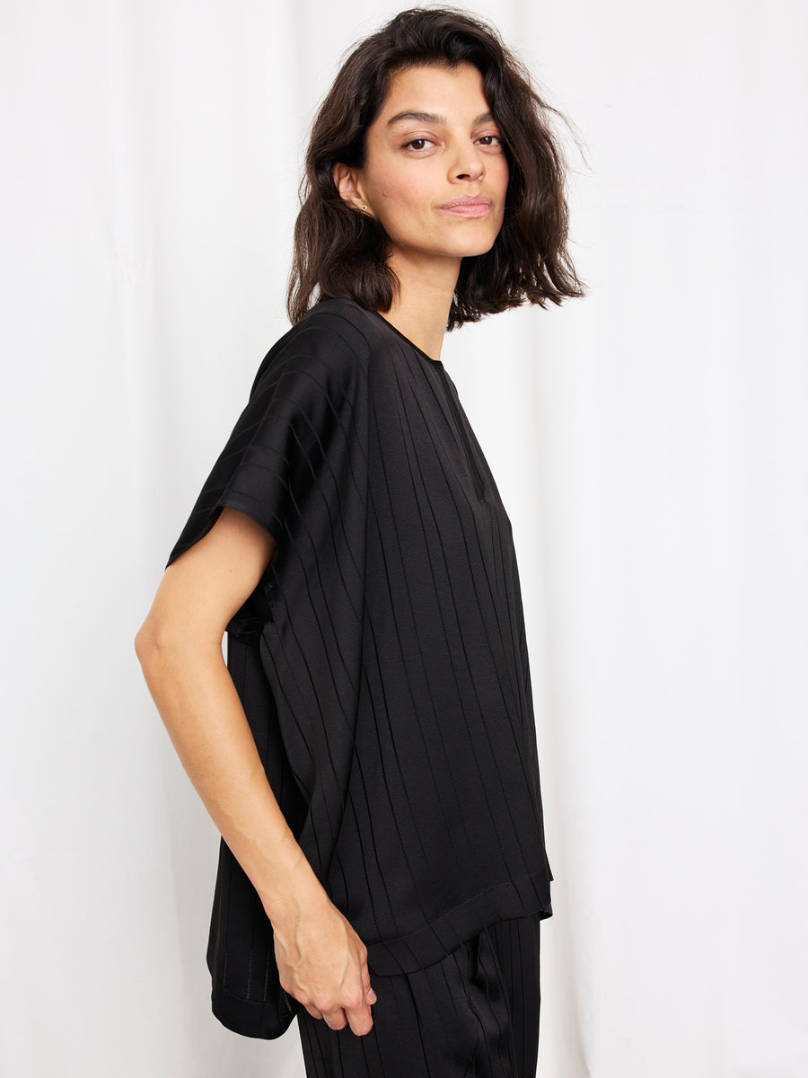 A woman with medium-length dark hair models the Rae Poncho Top by Zero + Maria Cornejo, standing sideways against a plain white background and looking confidently at the camera. This chic piece is made in New York.