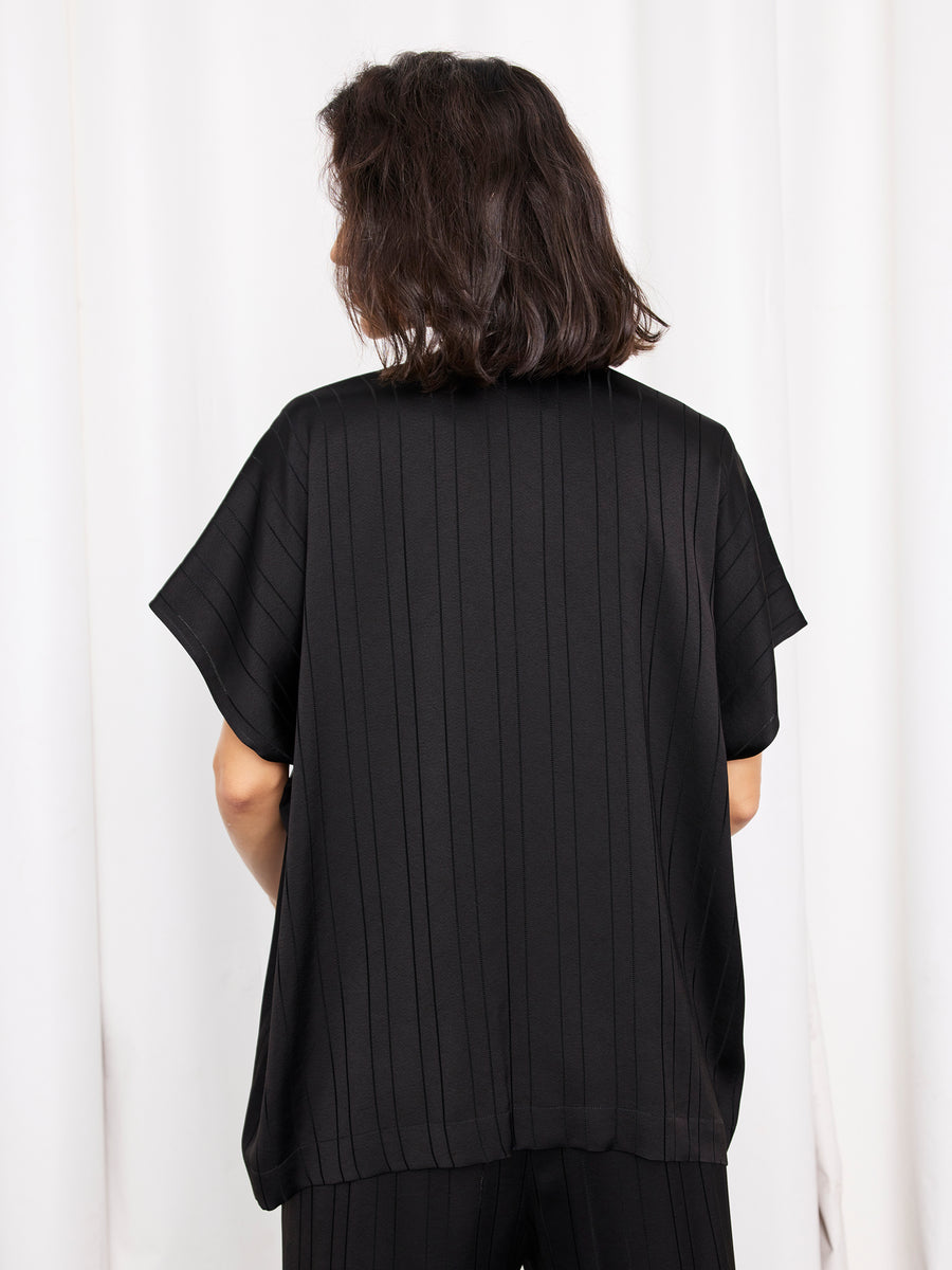 A person with shoulder-length dark hair, seen from behind, wears the Rae Poncho Top by Zero + Maria Cornejo—a woven striped piece with subtle vertical lines. The background is white curtains. This elegant top is made in New York.